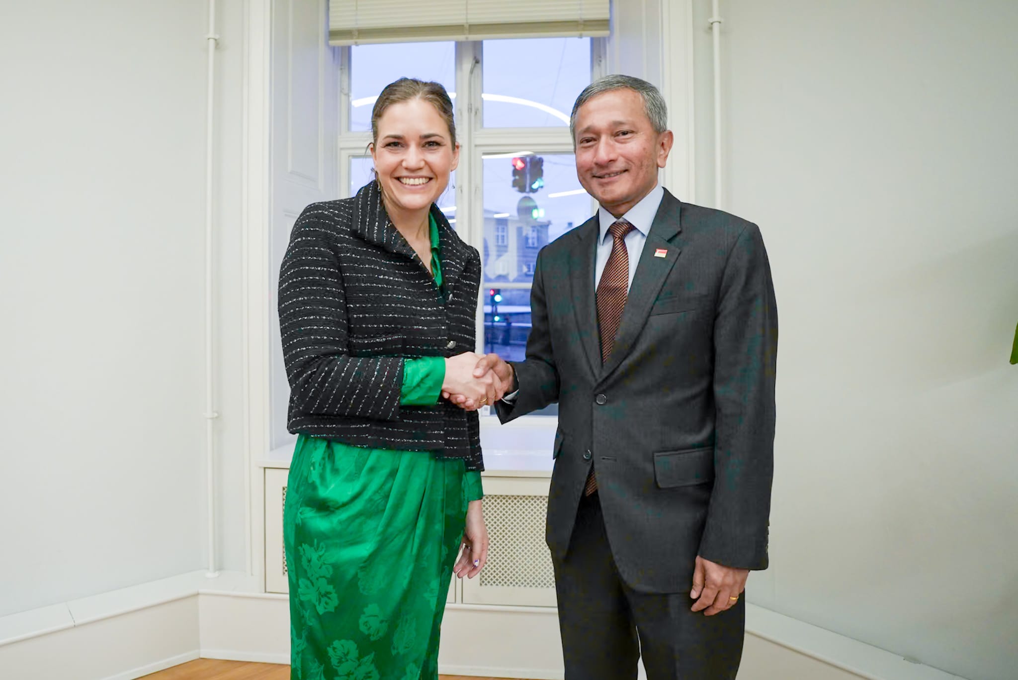 Two people shaking hands: a woman in green dress & jacket and a man in a gray suit and tie.